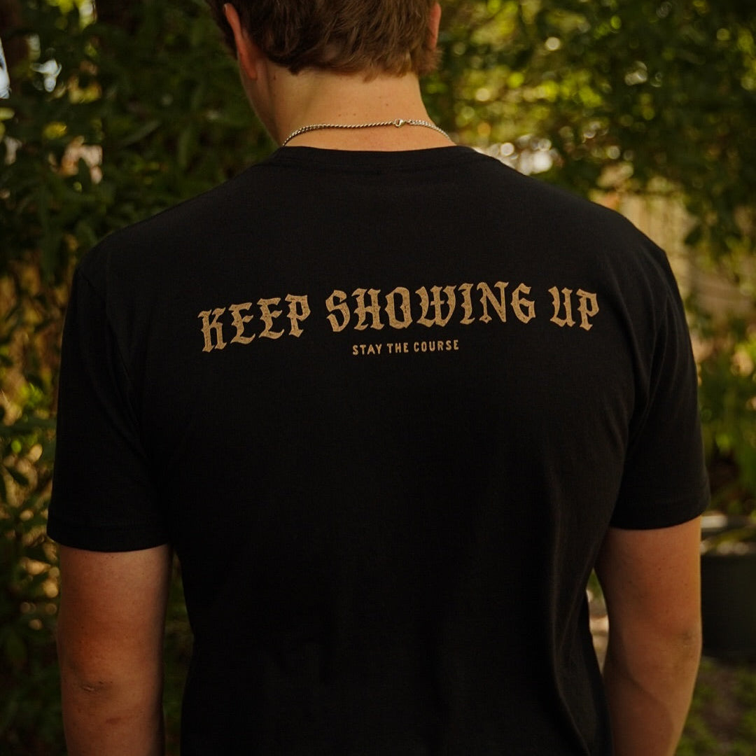 A person wearing a black T-shirt with white print on the back of a snake wrapped around a spear, with the words “ACTION OPENS DOORS” and “STAY THE COURSE”, set against a blurred outdoor background