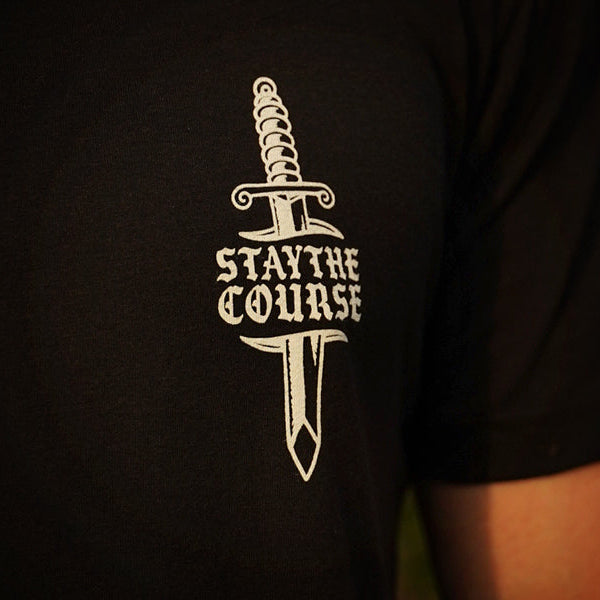 A close-up of a person wearing a black t-shirt with a white graphic of a dagger and the text 'STAY THE COURSE' printed on the chest.
