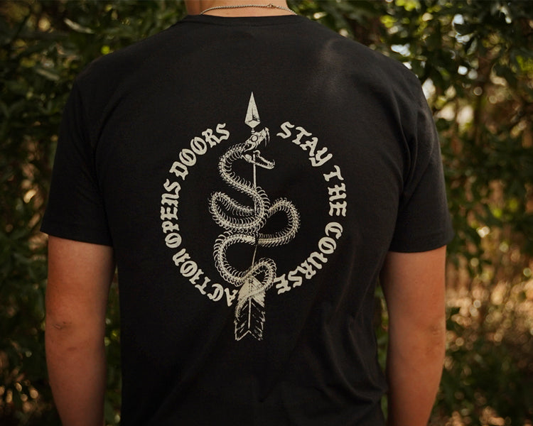 A person viewed from behind wearing a black short-sleeve T-shirt with a large white graphic print on the back. The design shows a coiled snake wrapped around a vertical spear, with the words “ACTION OPENS DOORS” on the left and “STAY THE COURSE” on the right, set against a blurred outdoor background of green foliage.