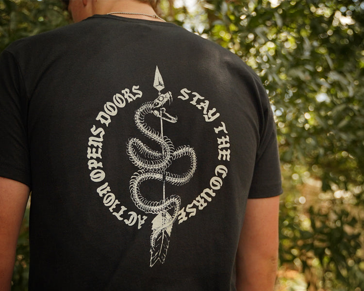 A person viewed from behind wearing a black T-shirt with a large white snake graphic and the text 'ACTION OPEN DOORS STAY THE COURSE' on the back, standing outdoors in front of blurred green foliage.