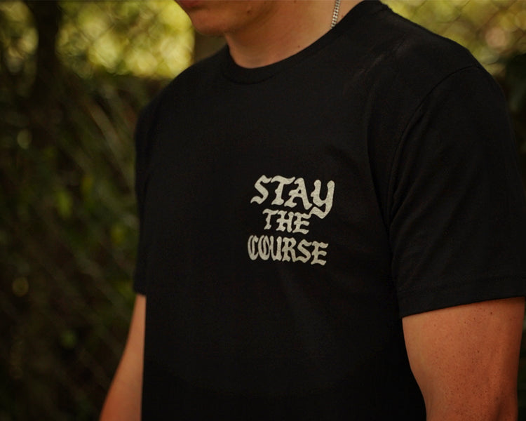 A person wearing a black t-shirt with white graphic that says STAY THE COURSE, standing against a blurred background