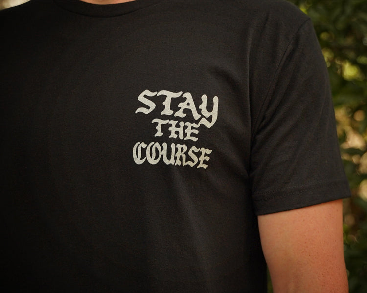 A person wearing a black t-shirt with white graphic that says STAY THE COURSE, standing against a blurred background