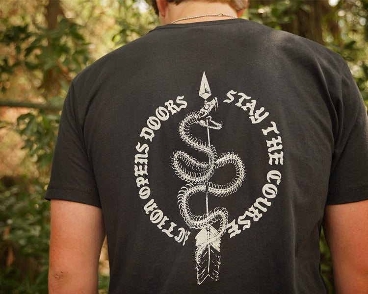 A person viewed from behind wearing a black T-shirt with a large white snake graphic and the text 'ACTION OPEN DOORS STAY THE COURSE' on the back, standing outdoors in front of blurred green foliage.