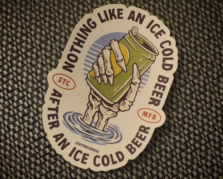 A die-cut sticker featuring a hand holding an ice-cold beer can above water, with the text 'NOTHING LIKE AN ICE COLD BEER AFTER AN ICE COLD BEER' around the edge, placed on a woven dark surface.