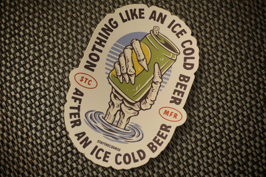 A die-cut sticker featuring a hand holding an ice-cold beer can above water, with the text 'NOTHING LIKE AN ICE COLD BEER AFTER AN ICE COLD BEER' around the edge, placed on a woven dark surface.