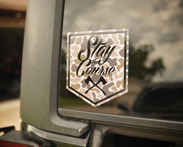 Camouflage-style 'Stay the Course' decal on a vehicle window with a blurred outdoor background.