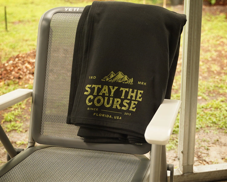 A black fleece blanket draped over a gray patio chair, featuring a green graphic that reads 'STAY THE COURSE' with mountains and 'FLORIDA, USA' on a porch with a grassy yard in the background.