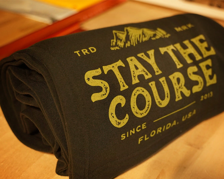 A rolled black blanket with distressed green text reading 'STAY THE COURSE', photographed on a wooden surface.