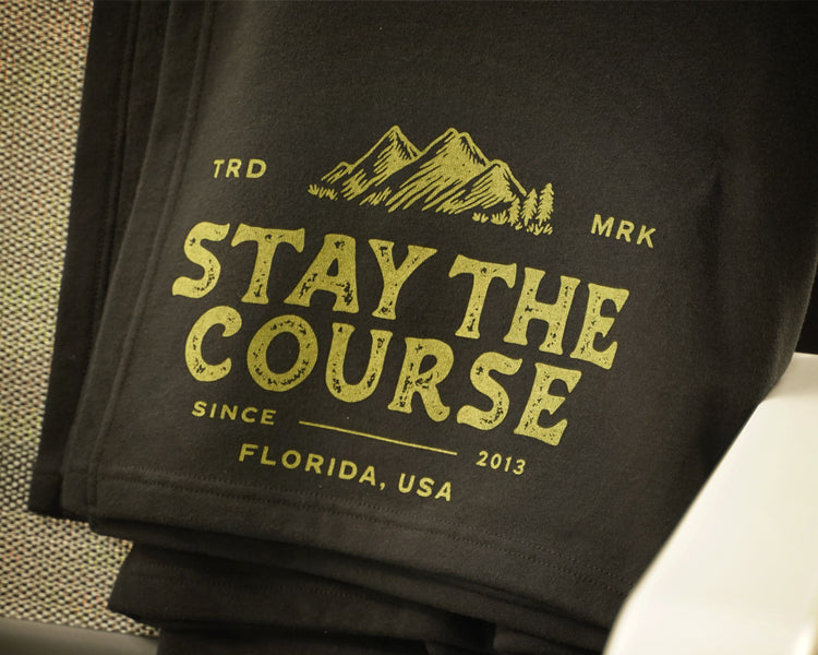 A black fleece blanket draped over a gray patio chair, featuring a green graphic that reads 'STAY THE COURSE' with mountains and 'FLORIDA, USA' on a porch with a grassy yard in the background.