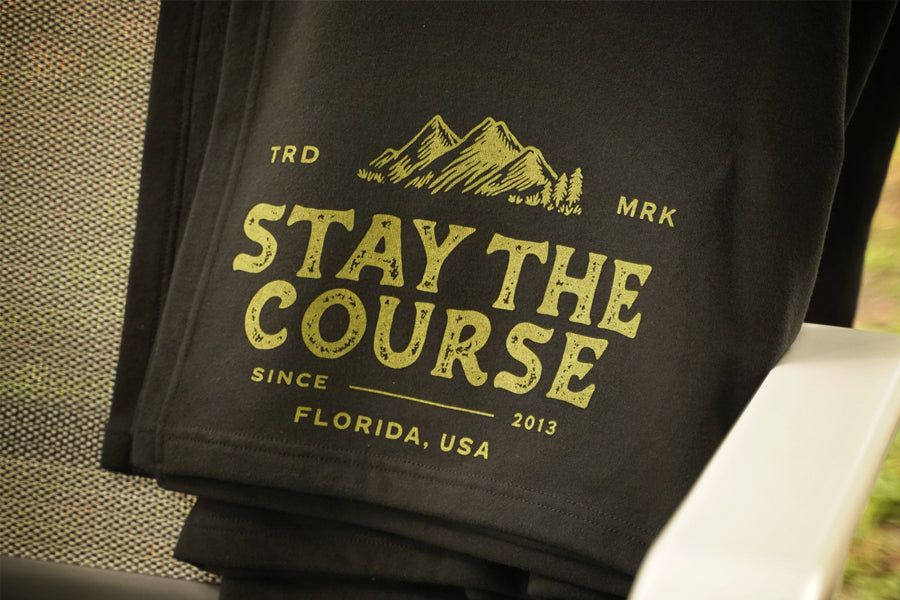 A black fleece blanket draped over a gray patio chair, featuring a green graphic that reads 'STAY THE COURSE' with mountains and 'FLORIDA, USA' on a porch with a grassy yard in the background.