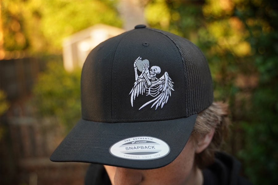 Black snapback trucker cap with skeleton design worn by a person, blurred background