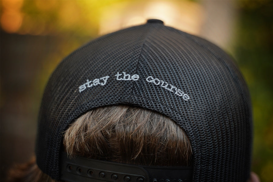 Black cap with 'stay the course' text worn by a person, blurred natural background