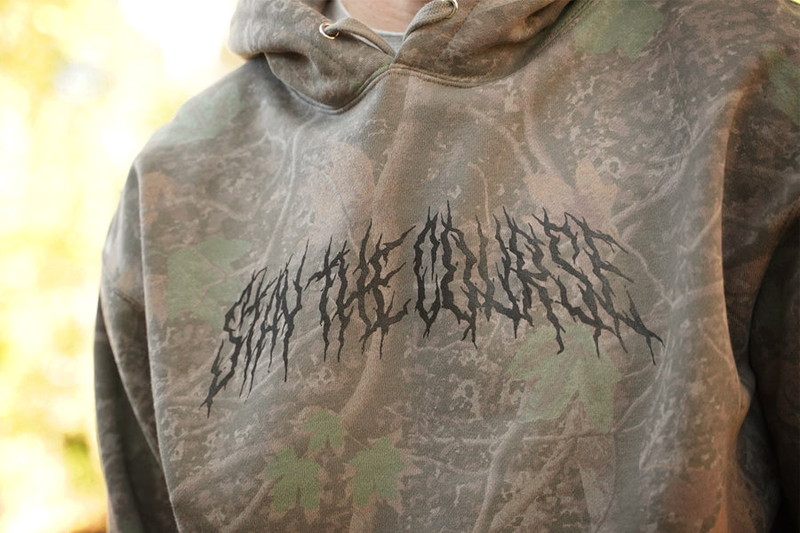 Camouflage hoodie with black stay the course text design worn by a person, blurred background