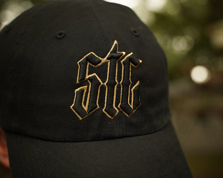 Black cap with gold embroidered logo against a blurred natural background