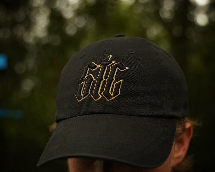 Black cap with STC black and gold logo worn by a person, blurred background