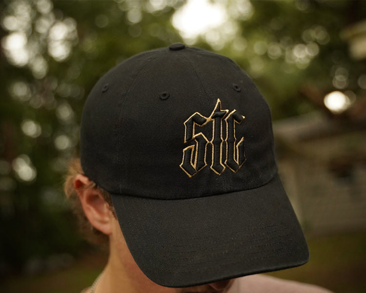 Black cap with gold logo worn by a person outdoors