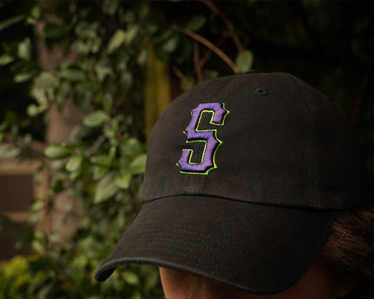 Person wearing a black cap with a purple 'S' logo against a blurred natural background
