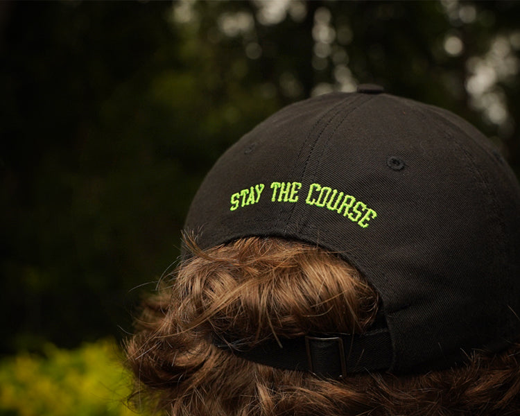 Black cap with 'Stay the Course' text worn by a person outdoors