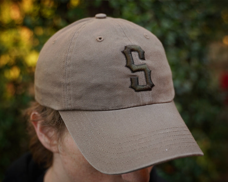 Beige cap with a logo worn by a person against a blurred natural background