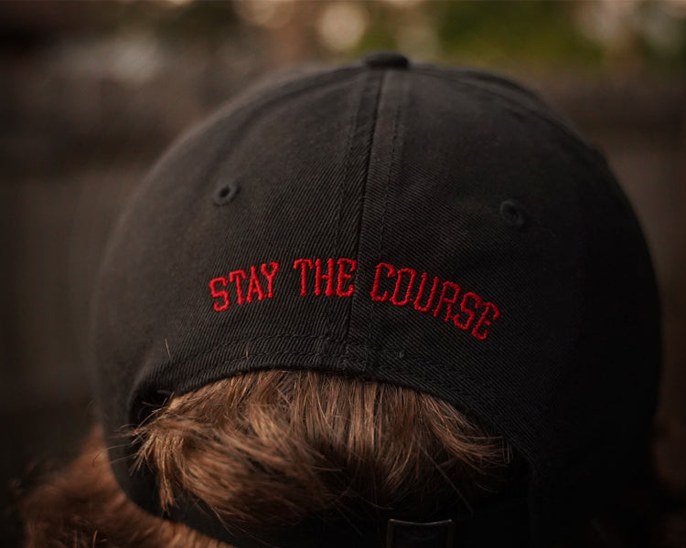 Black cap with 'Stay the Course' text worn by a person, blurred background