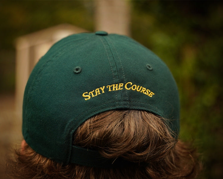 Green cap with 'Stay the Course' text worn by a person, blurred background