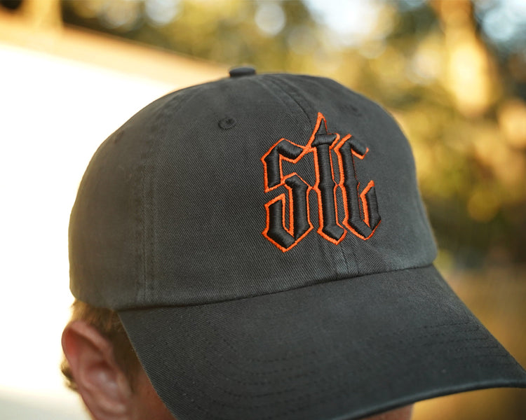 Person wearing a black hat with orange and black "stc" puff embroidery on the front, in an outdoor setting