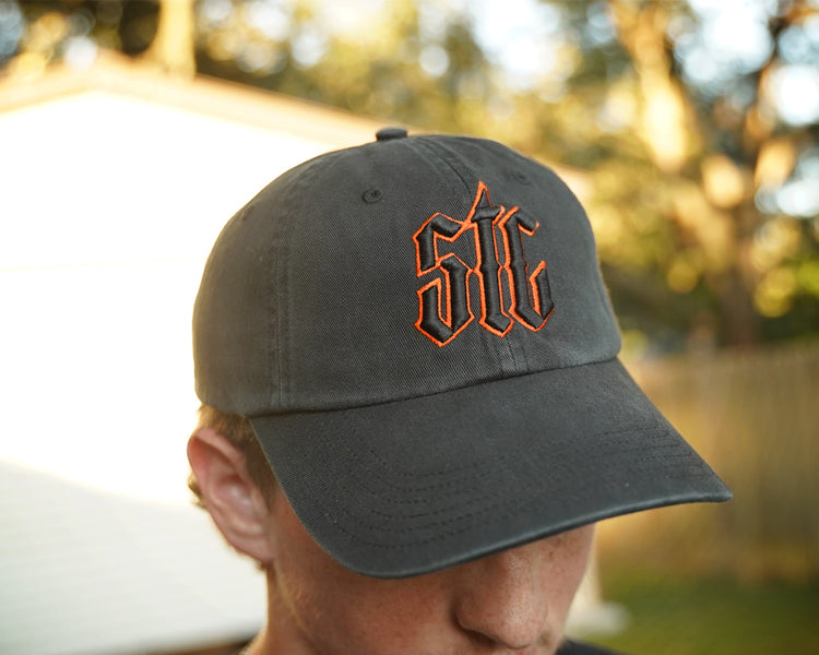Person wearing a black hat with orange and black "stc" puff embroidery on the front, in an outdoor setting