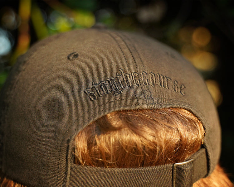 Person wearing a black hat with black "stay the course" embroidery on the back, in an outdoor setting