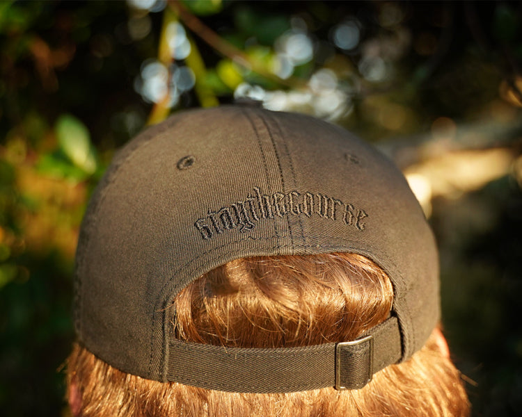 Person wearing a black hat with black "stay the course" embroidery on the back, in an outdoor setting