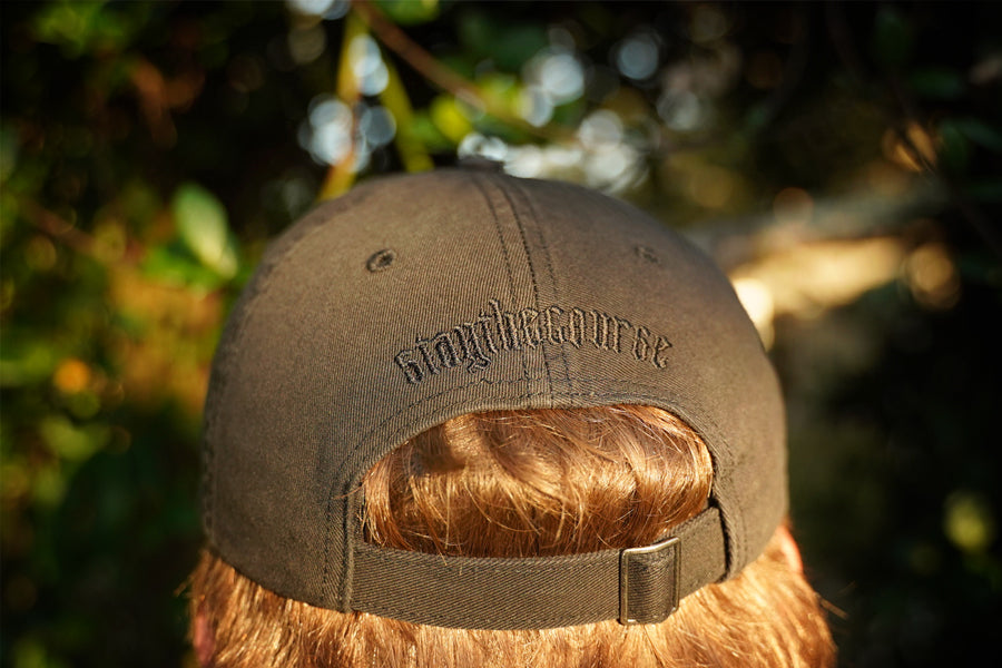 Person wearing a black hat with black "stay the course" embroidery on the back, in an outdoor setting