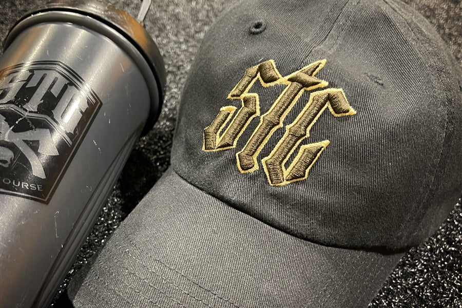 Black cap with gold and brown logo and black blender bottle on a textured surface