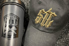 Black tumbler and black cap with gold and brown logo on a textured surface