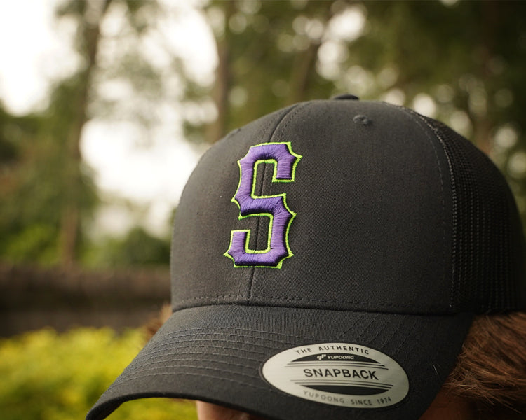 Black trucker cap with a purple and green logo worn by a person outdoors.