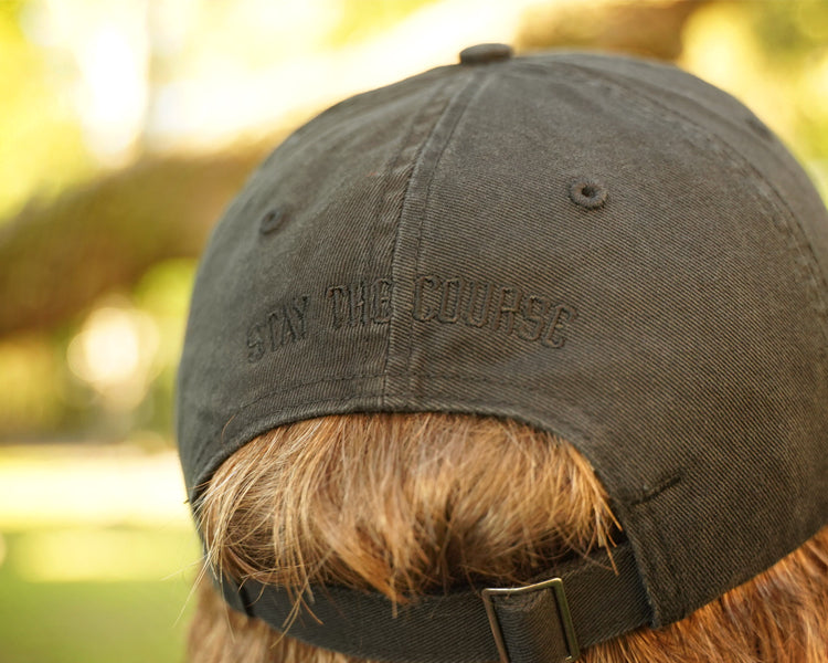 Person wearing a black hat with black embroidery on the back that says "stay the Course", in an outdoor setting