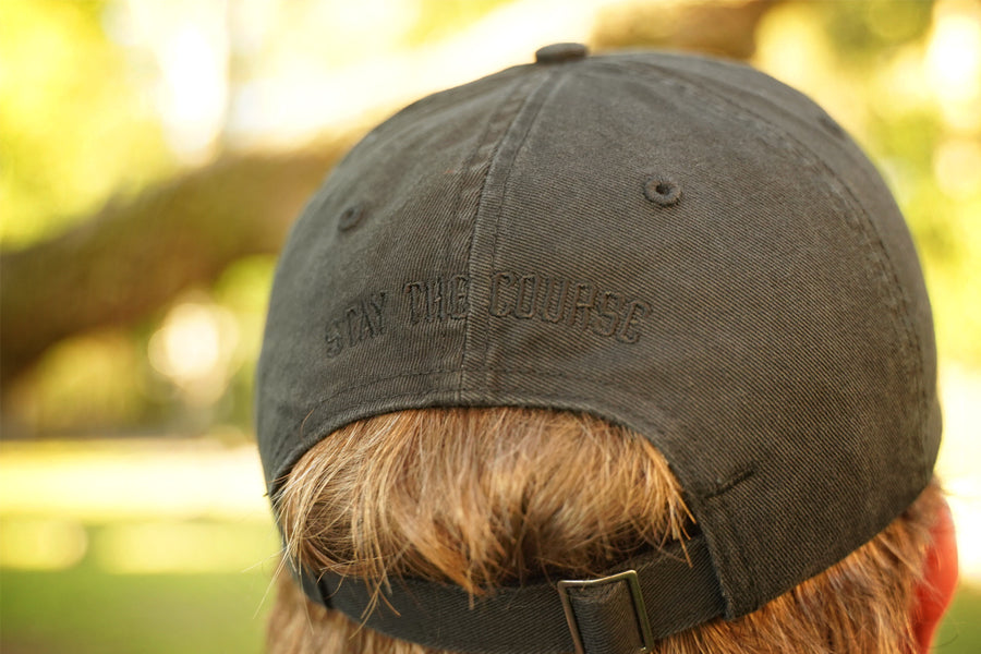 Person wearing a black hat with black embroidery on the back that says "stay the Course", in an outdoor setting
