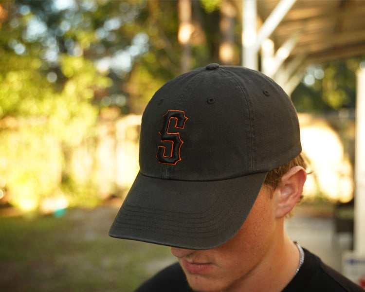 Person wearing a black hat with orange and black "s" puff embroidery on the front, in an outdoor setting