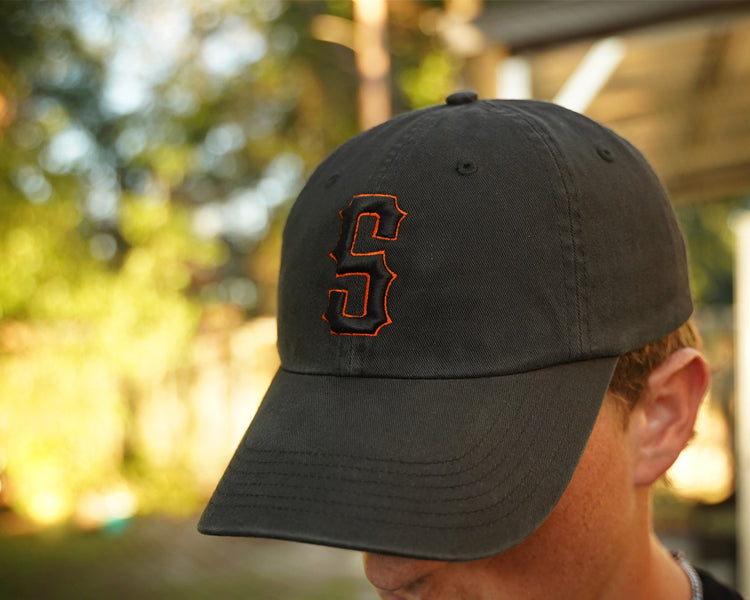 Person wearing a black hat with orange and black "s" puff embroidery on the front, in an outdoor setting