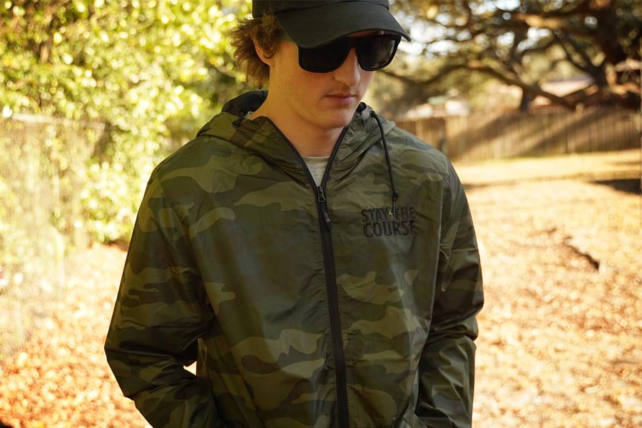 Person wearing a camouflage jacket with 'Stay the course' branding outdoors.