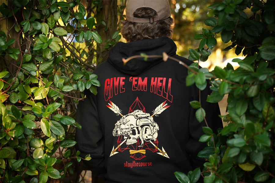 Person wearing a black hoodie with a graphic design and text, standing among green foliage.