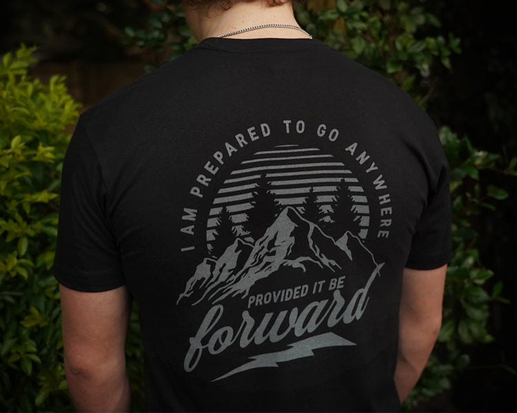 Person wearing a black t-shirt with a mountain design and text on the back, standing against a green leafy background.