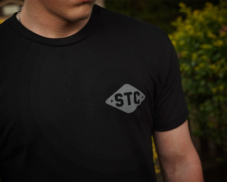 Person wearing a black t-shirt with 'STC' logo against a blurred green background