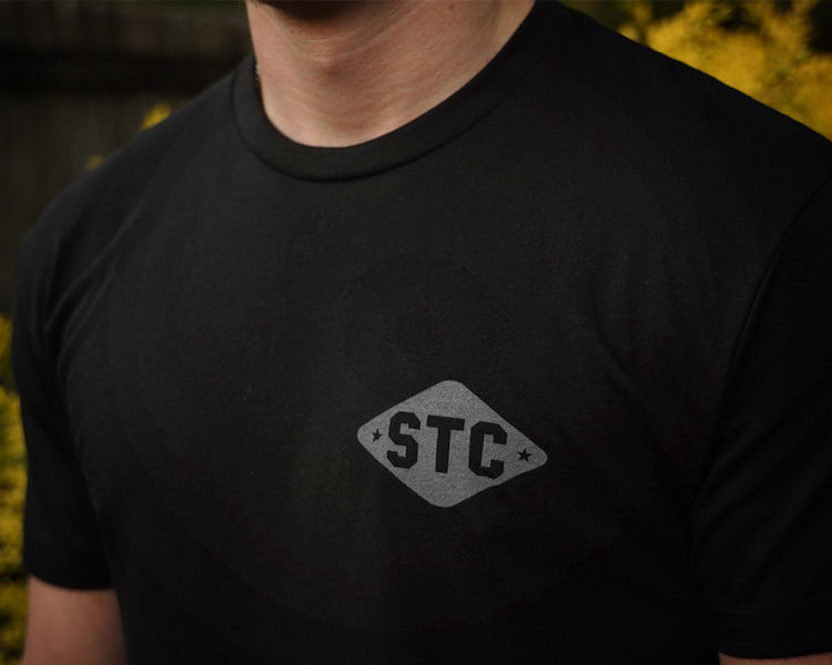 Person wearing a black t-shirt with 'STC' logo against a blurred natural background