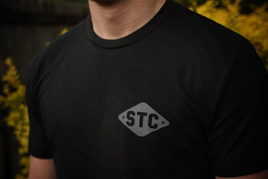 Person wearing a black t-shirt with 'STC' logo against a blurred natural background