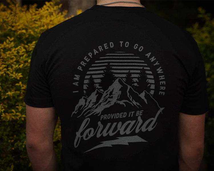 Person wearing a black t-shirt with a mountain design and text on the back, standing in front of greenery.