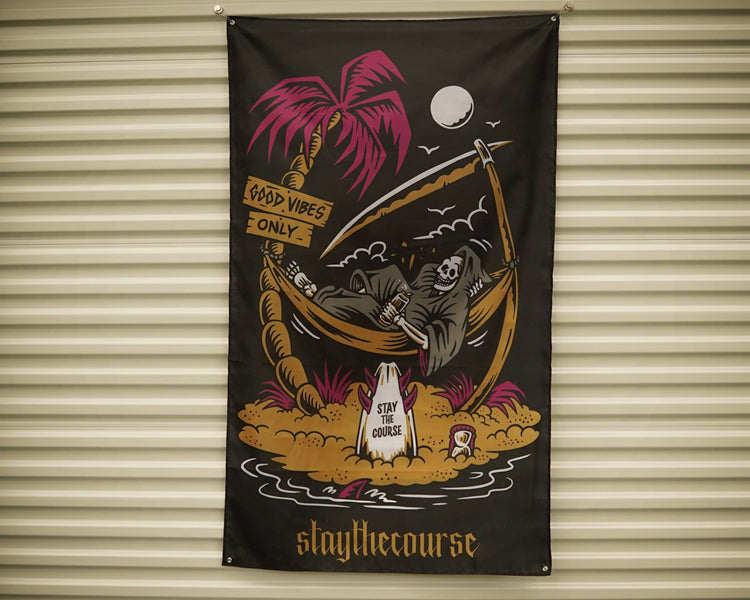A black fabric banner featuring a colorful graphic of a skeleton reclining in a palm tree hammock on a tropical island, with the text 'staythecourse' at the bottom, hanging against a beige corrugated metal wall.