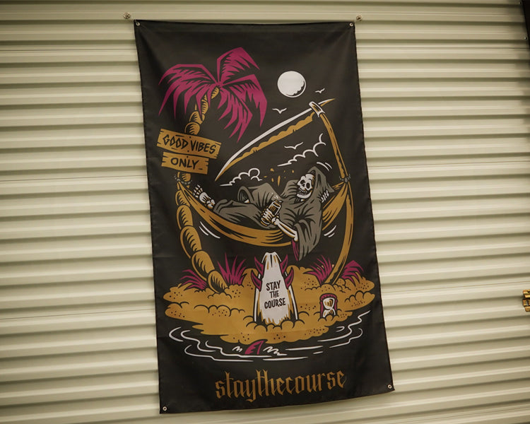 A black vertical banner featuring a grim reaper lounging in a palm tree hammock on a beach, with a pink palm tree, moon, and the text 'staythecourse' at the bottom, hanging against a beige corrugated metal wall.