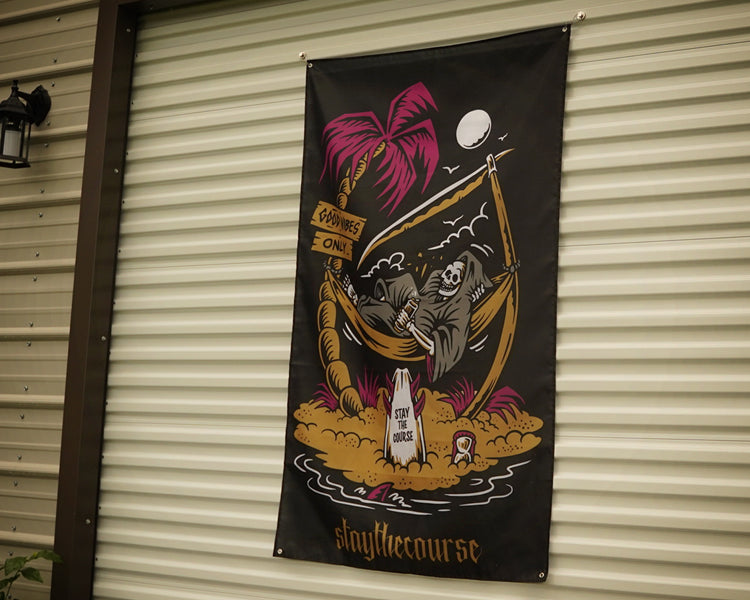 A vertical black banner hanging on a beige corrugated metal garage door. The banner features a colorful graphic illustration of a skeleton in a hammock under a palm tree, with the text 'slaythe course' at the bottom.