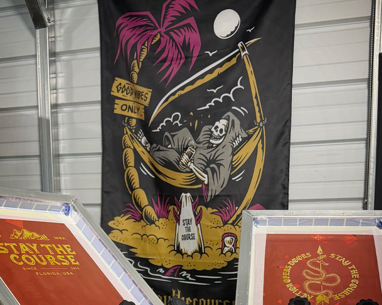 A large black fabric banner featuring a colorful graphic of a skeleton reclining in a hammock under a palm tree, displayed indoors in front of a white siding wall. Two framed prints are partially visible in the foreground.