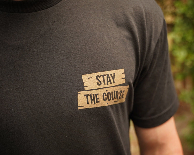 Black t-shirt with 'Stay The Course' text on a blurred natural background