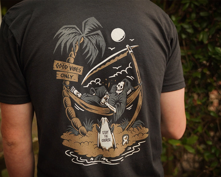 Person wearing a black t-shirt with a graphic design of a skeleton in a hammock, palm trees, and text.